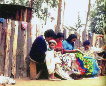 The Collective Embroidery Cuatro mujeres bordando el Bordado Colectivo junto a un niño pequeño frente a un muro de tablas en Isla Negra